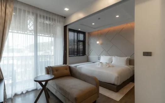 Спальня с белым постельным бельём и бежевым шезлонгом у окна