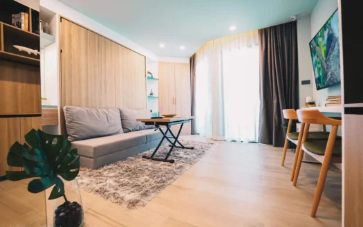 Яркая гостиная с серым диваном, шкафами из светлого дерева и большим окном